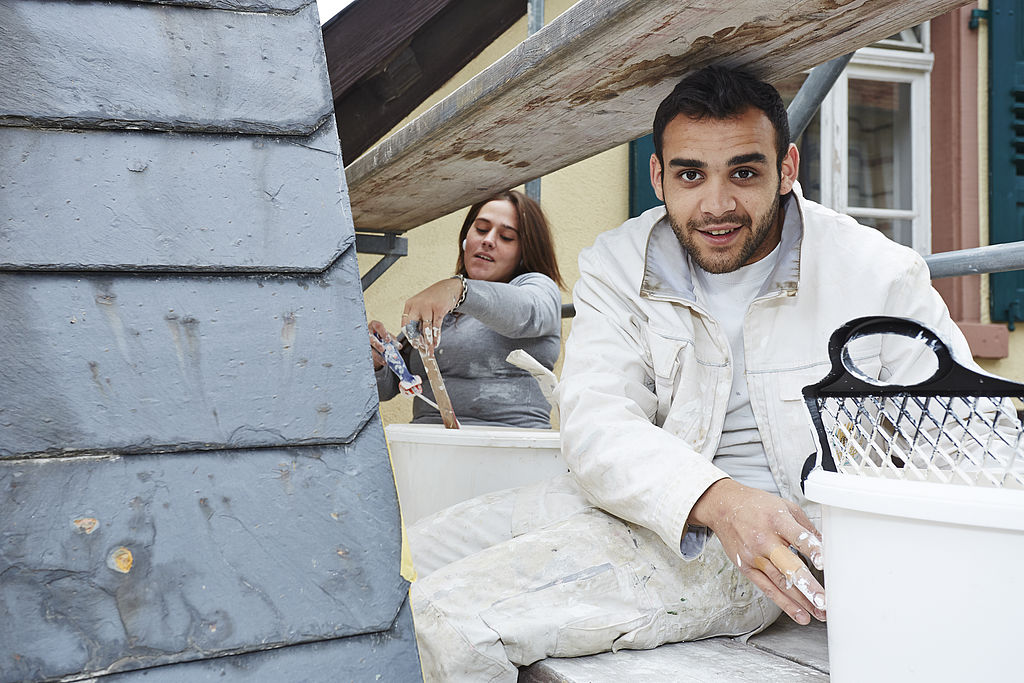 Zwei Azubis auf der Baustelle, eine junge Frau und ein junger Mann auf einem Gerüst an der Außenfassade eines Hauses